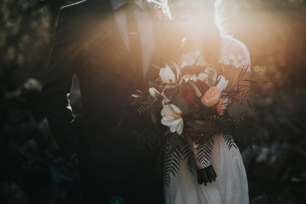 A couple standing close together, the groom in a dark suit and the bride in a white dress, holding a vibrant bouquet of flowers including roses and lilies, with sunlight creating a warm glow around them.
