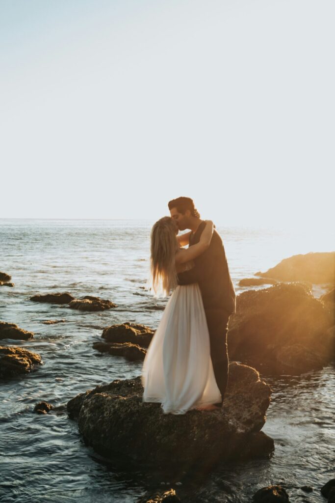 A couple embracing and kissing on rocky beach at sunset, with golden sunlight illuminating the scene and the ocean in the background.