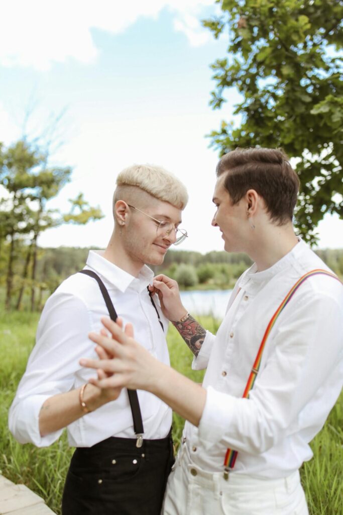 Two individuals with stylish haircuts and attire, standing close together in a green outdoor setting, sharing a tender moment. One is adjusting the other's suspenders while holding hands.