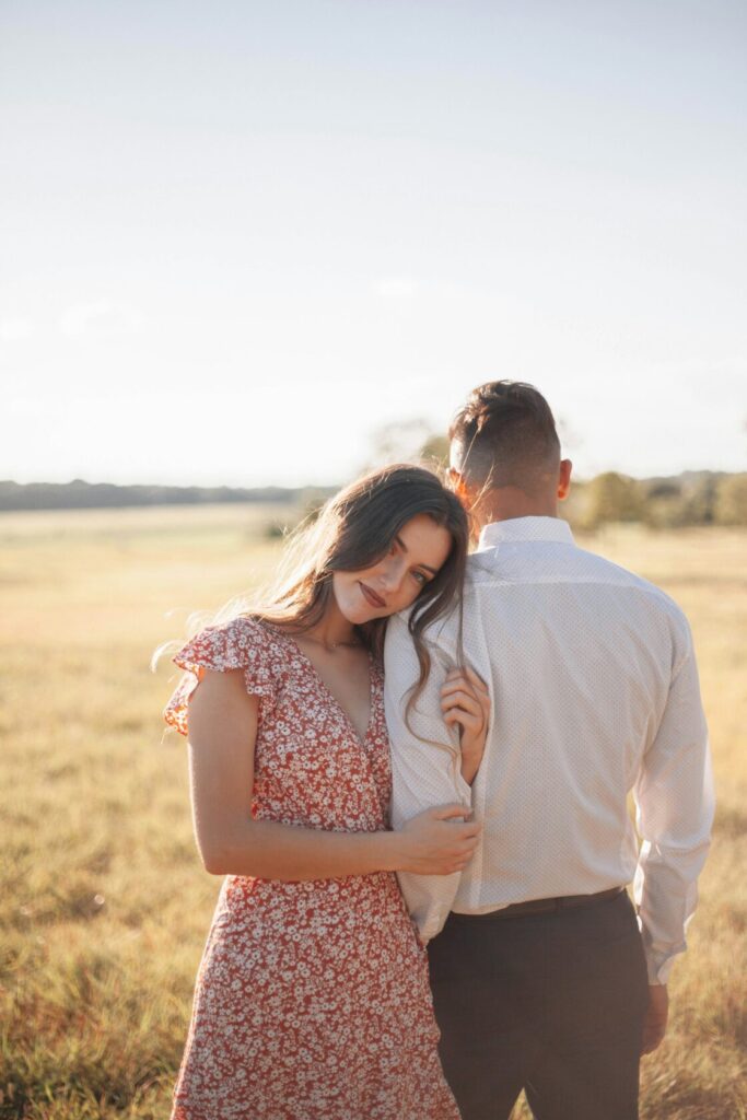A young woman in a floral dress leans affectionately against a man in a white shirt, both standing in a sunlit field with tall grass and a clear sky.