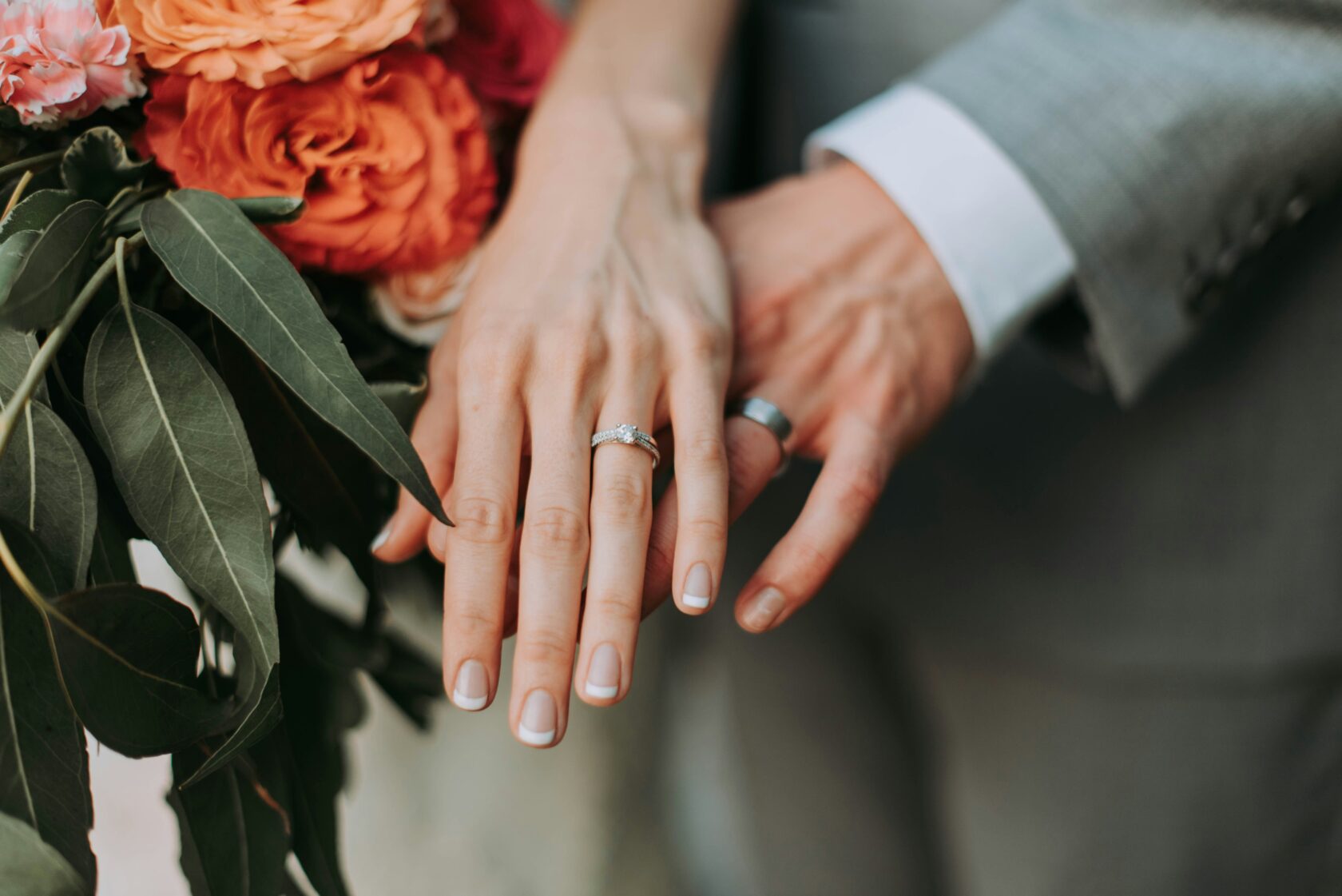 Close-up of a couple's hands, showcasing wedding rings on the left hand and a beautiful floral bouquet in the background.