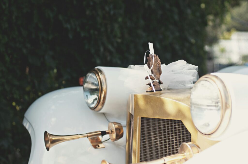 Close-up of a white vintage car's bonnet decorated with a brown eagle ornament and white tulle fabric, featuring brass horn embellishments and round headlights.