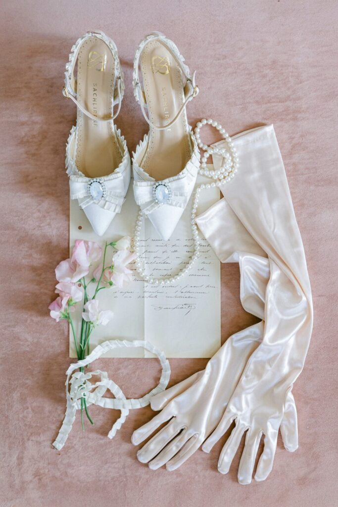 A pair of white bridal shoes adorned with a bow and gemstones, accompanied by satin gloves, pearl necklaces, delicate flowers, and a vintage letter on a soft pink velvet surface.