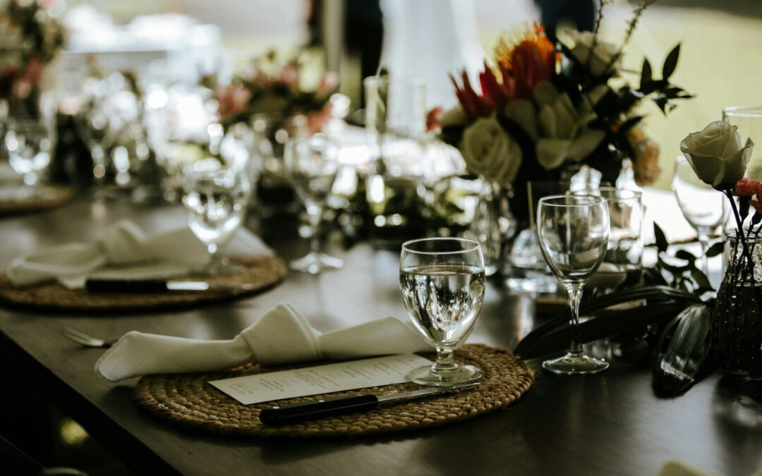 A beautifully arranged dining table with woven placemats, elegant glassware, folded napkins, cutlery, and floral centrepieces.