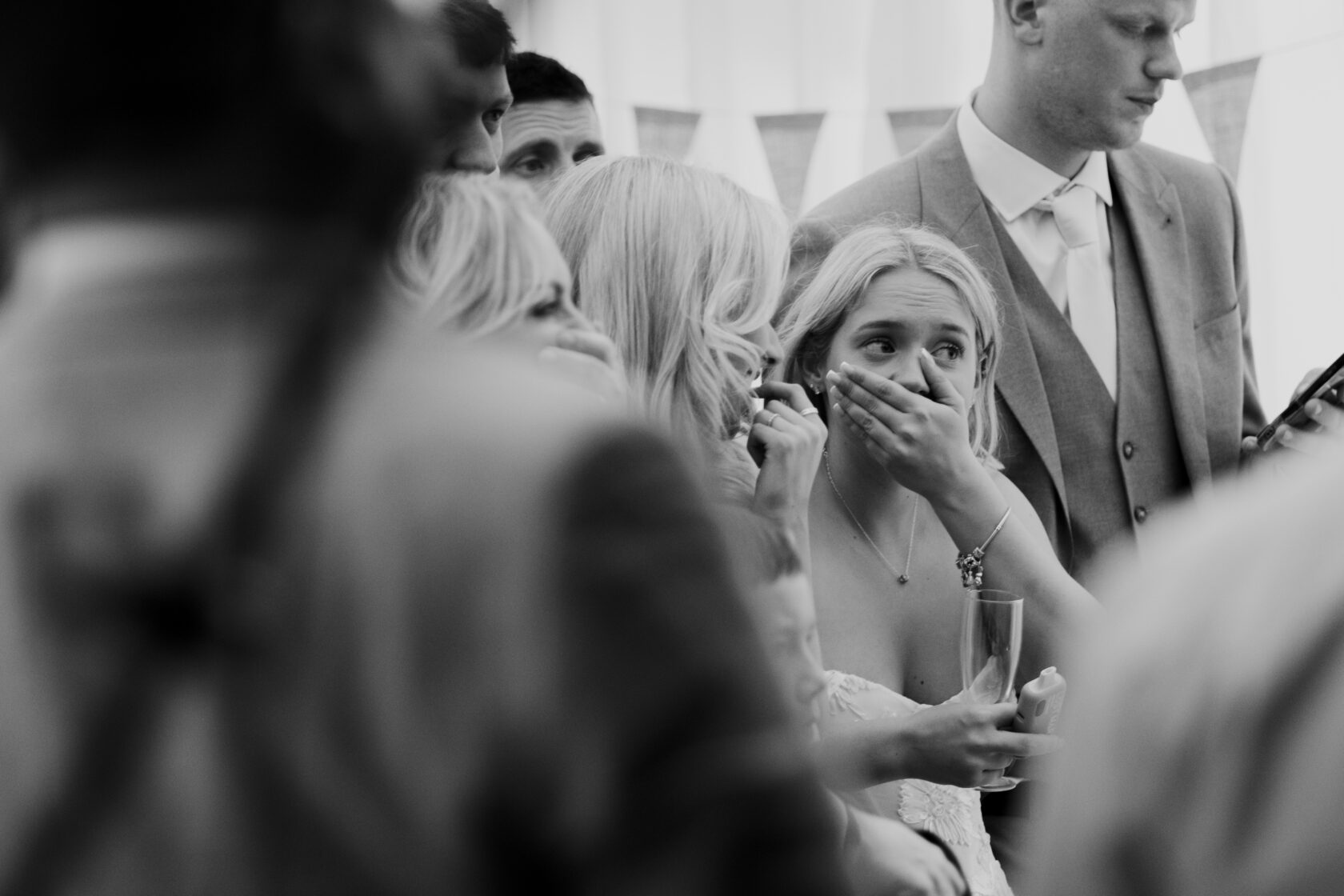 A close-up of several guests at a wedding displaying emotional reactions. A bride, with her hands covering her mouth, appears to be captivated by an unexpected moment, while others around her exhibit similar expressions. Shot for Lydia Turner.