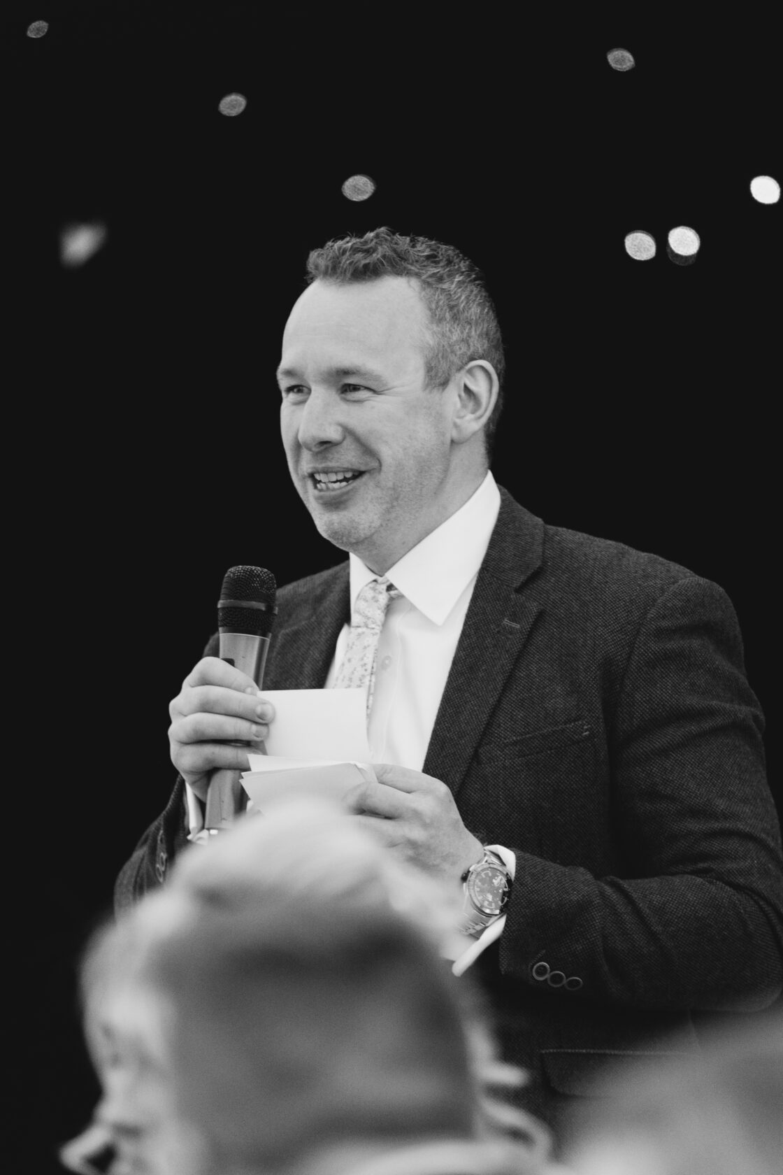 A smiling man holding a microphone and notes, delivering a speech at an event. He is dressed in a suit, engaging with the audience. Shot for Lydia Turner.
