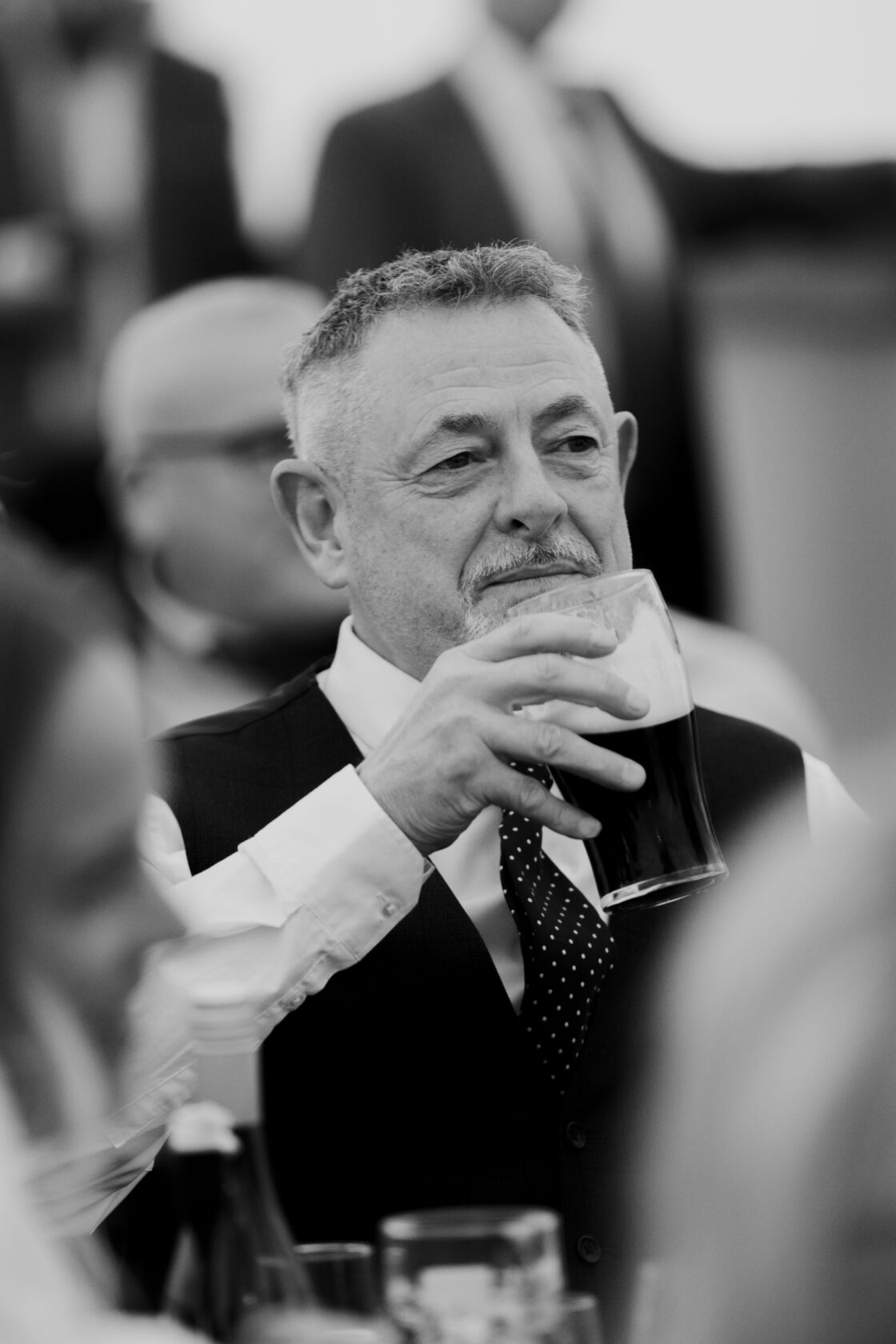 A middle-aged man with grey hair wearing a black vest and a white shirt with a polka dot tie, holding a pint of dark beer, smiling at an event. Shot for Lydia Turner