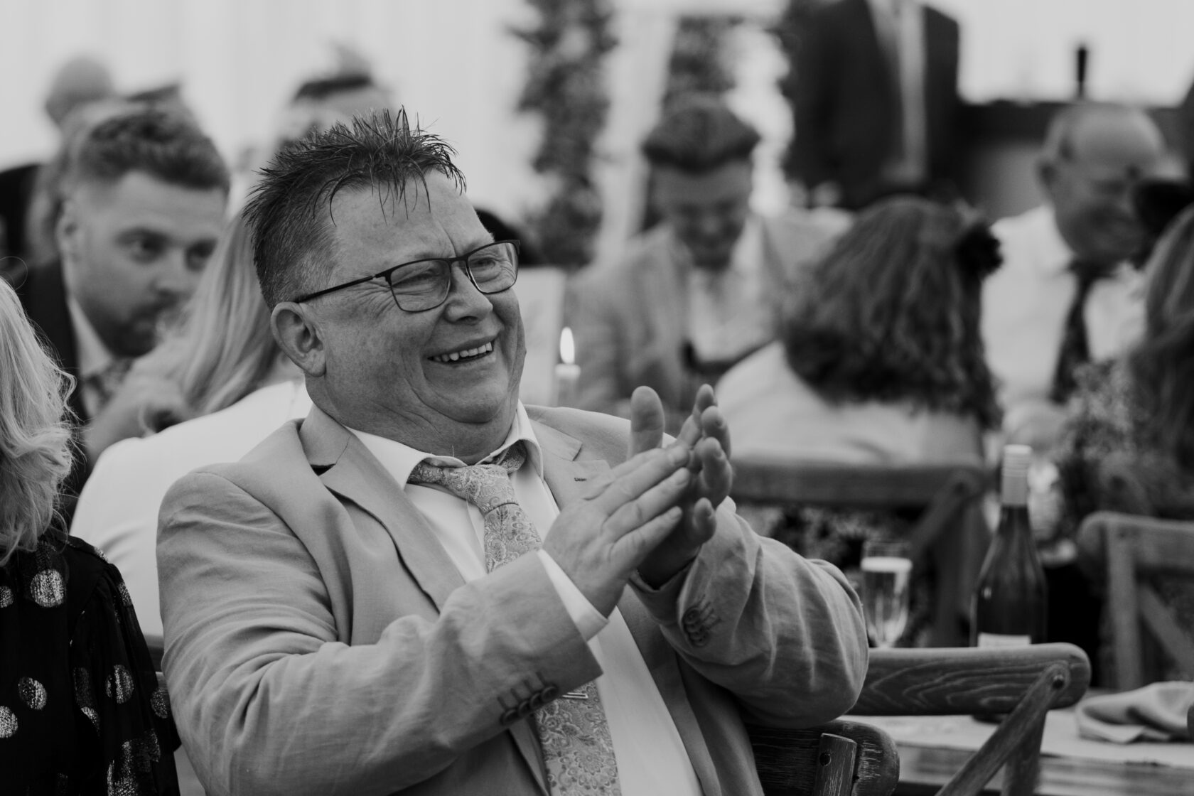 A man in a light grey suit and glasses laughing and clapping at a gathering, surrounded by happy guests in a festive atmosphere. Shot for Lydia Turner.