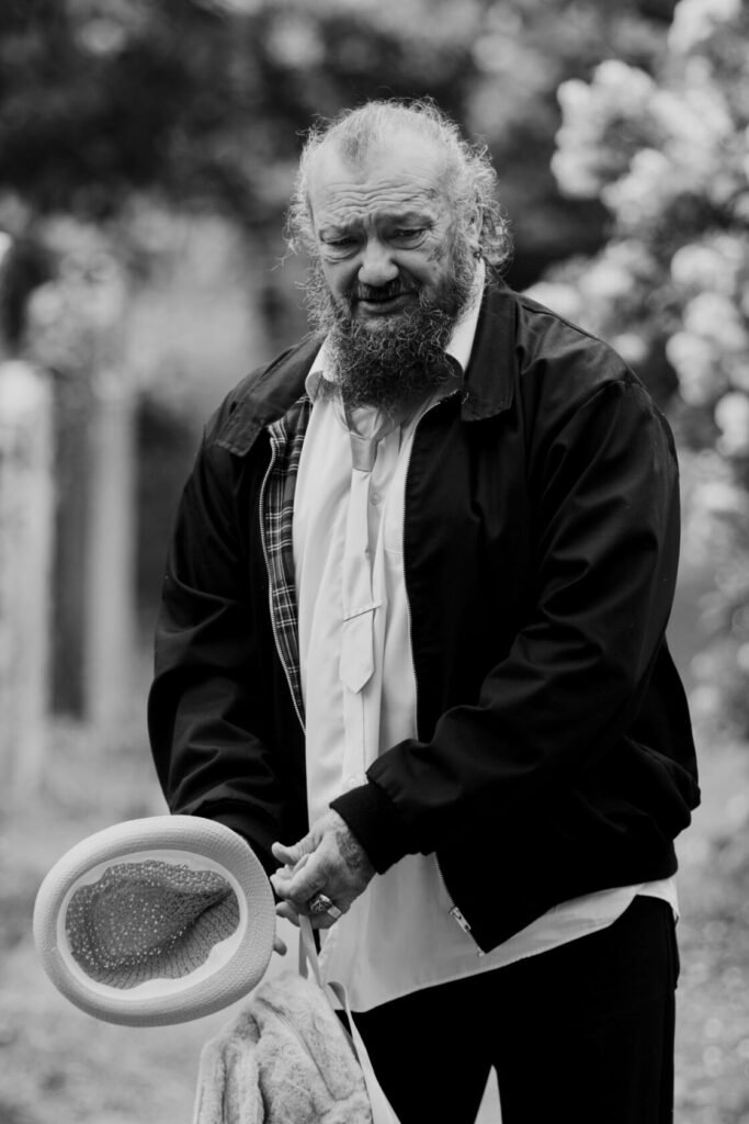 A bearded man dressed in a black jacket and white shirt holds a light-coloured hat, standing in a garden with blurred flowers in the background.