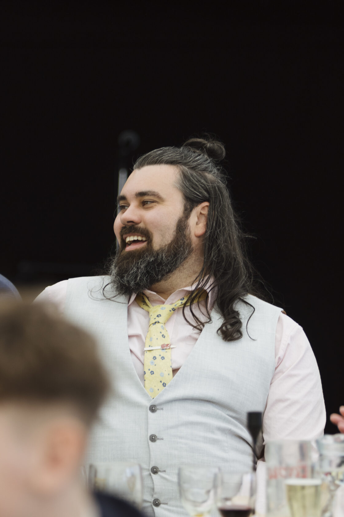 A smiling man with long hair and a beard, wearing a grey waistcoat and a floral tie, sitting at a table filled with drinks. Shot for Lydia Turner.