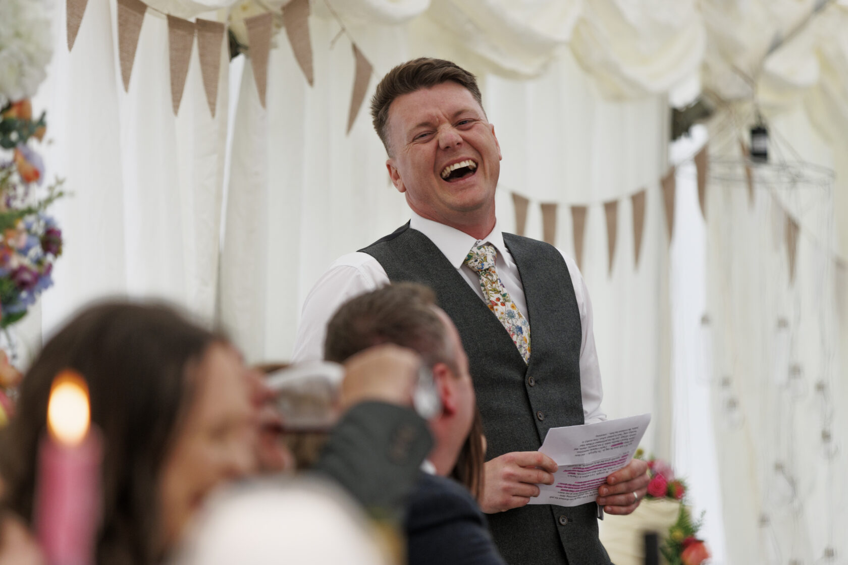 A smiling man in a grey waistcoat and floral tie delivers a speech at a wedding, with guests laughing and enjoying the moment. Shot for Lydia Turner.