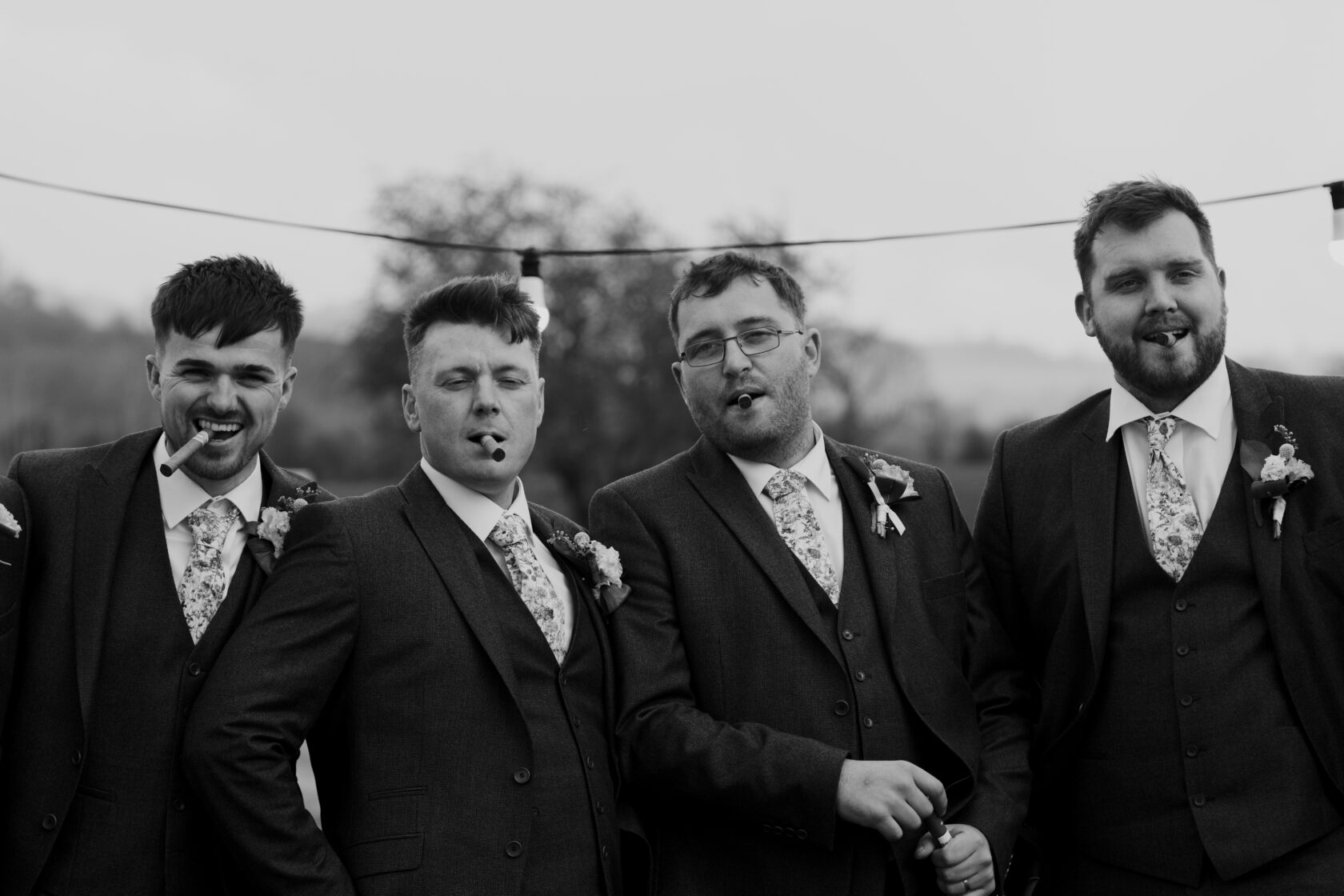 Four groomsmen in grey suits posing cheerfully with cigars in their mouths, adorned with floral ties and buttonholes. Shot for Lydia Turner.