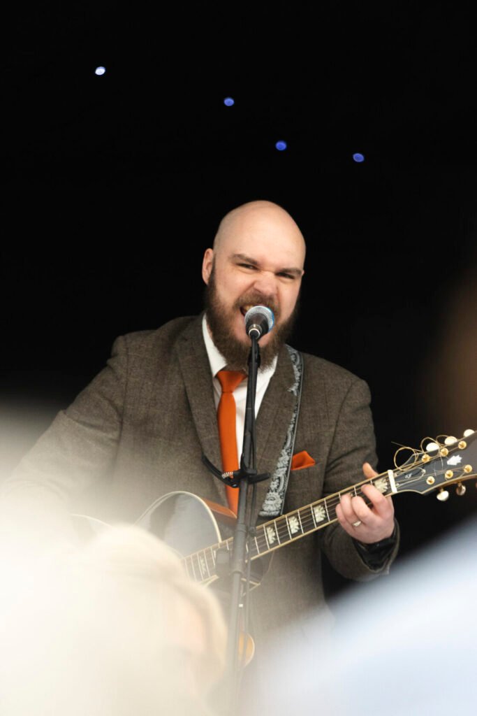 A bearded musician in a brown suit with an orange tie passionately performing on stage with a guitar, microphone in front, under a dark backdrop with twinkling lights.