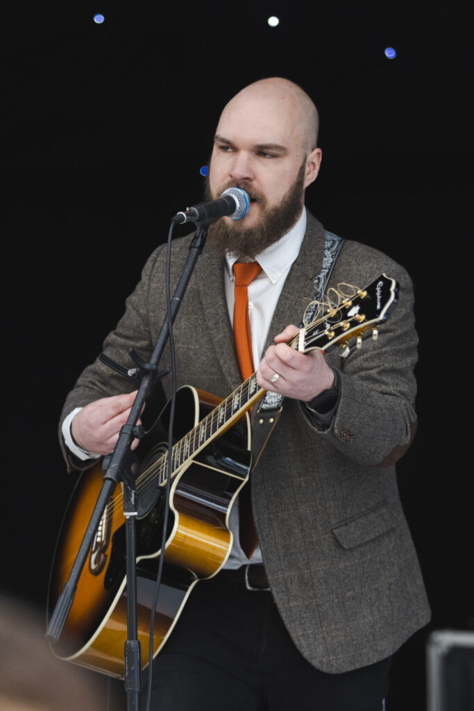 Singer-songwriter with a beard wearing a grey tweed jacket and orange tie, performing with an acoustic guitar at a microphone on stage.
