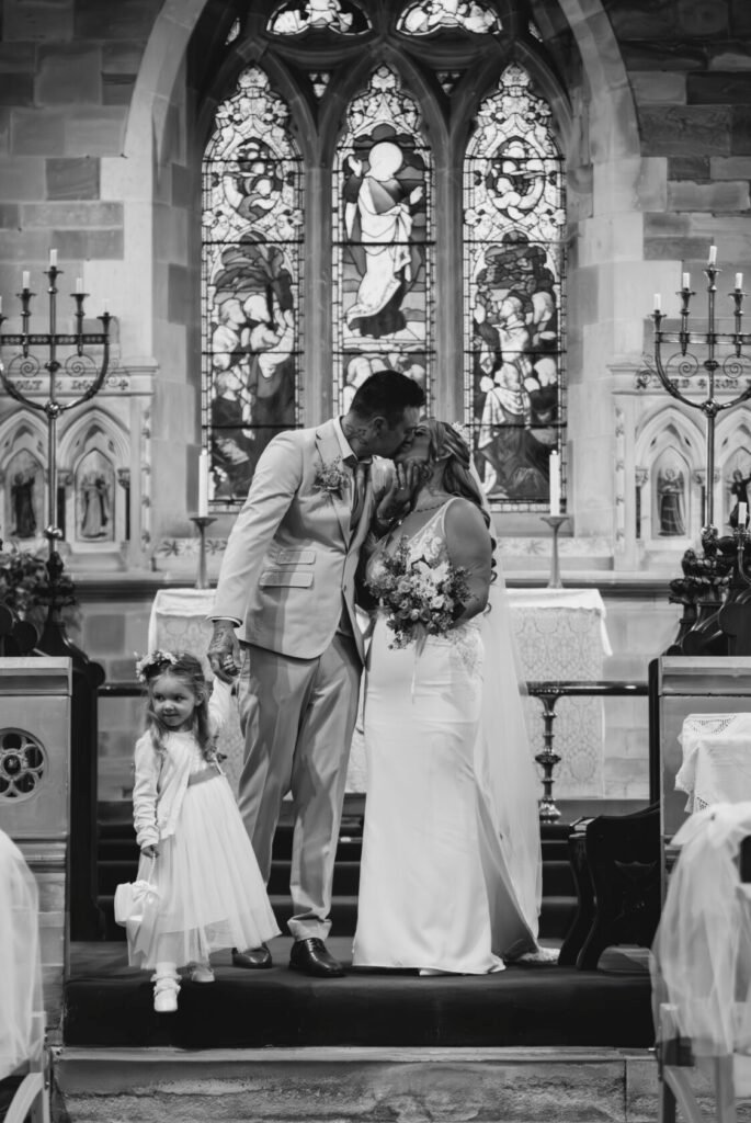 A bride and groom share a kiss during their wedding ceremony in a church, with stunning stained glass windows in the background. A young girl in a white dress stands beside them, holding their hands.