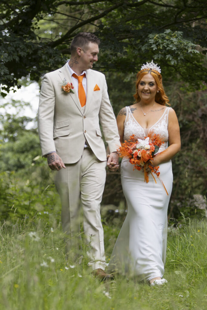 A bride and groom walking hand in hand through a green pathway. The bride, with orange hair and a floral bouquet in shades of orange, wears a fitted white gown, while the groom is dressed in a light-coloured suit with an orange tie.