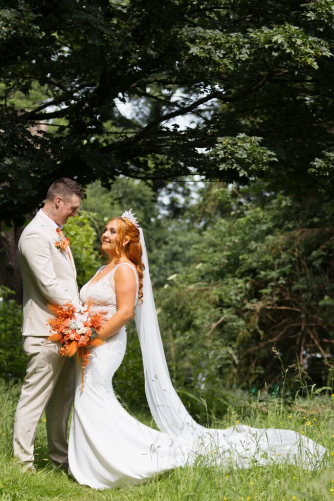 A bride and groom embrace in a lush green setting, with the bride wearing a white dress and holding a vibrant bouquet of orange flowers.
