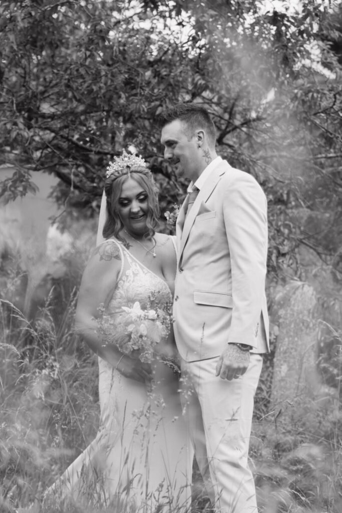 A bride and groom share a joyful moment outdoors, surrounded by tall grass and trees. The bride is adorned with a tiara and holding a bouquet, while the groom wears a light-coloured suit.
