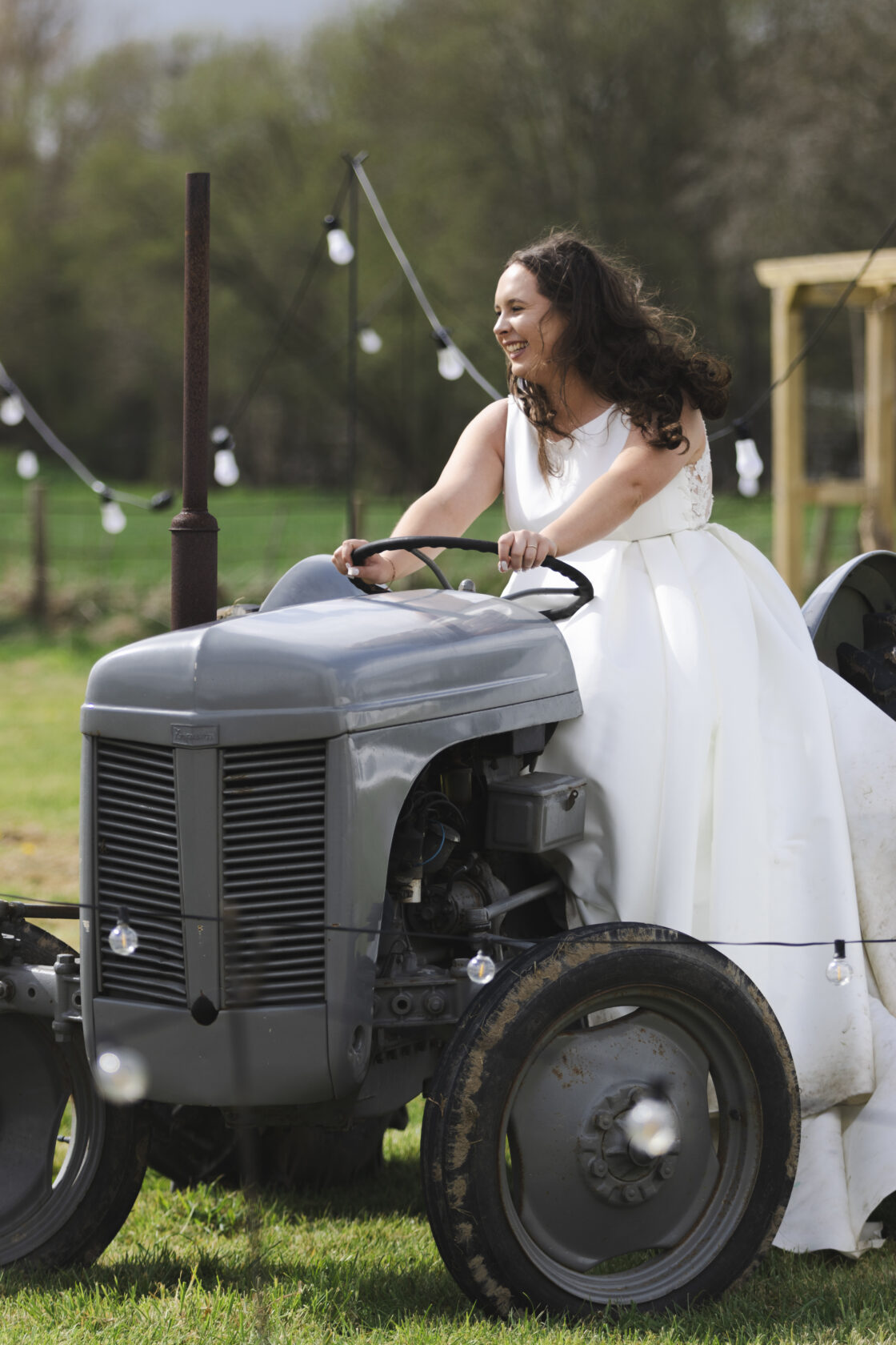 A joyful bride in a white wedding dress, laughing while driving a vintage grey tractor in a scenic outdoor setting with fairy lights in the background. Shot for Lydia Turner