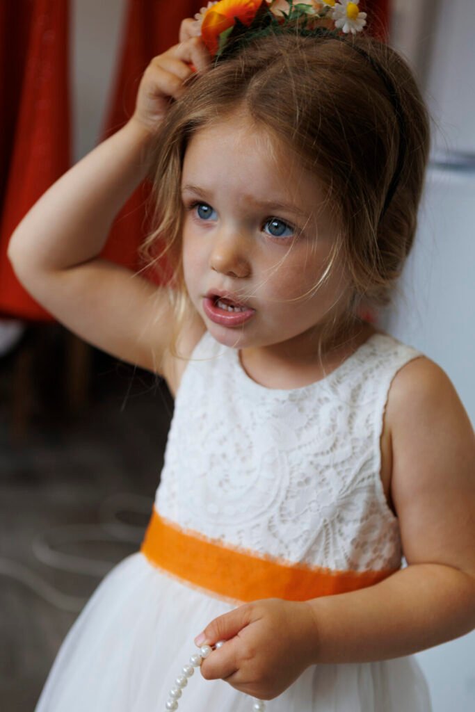 A young girl with blue eyes wearing a white lace dress adorned with an orange belt, adjusting a floral headband while holding a string of pearls.