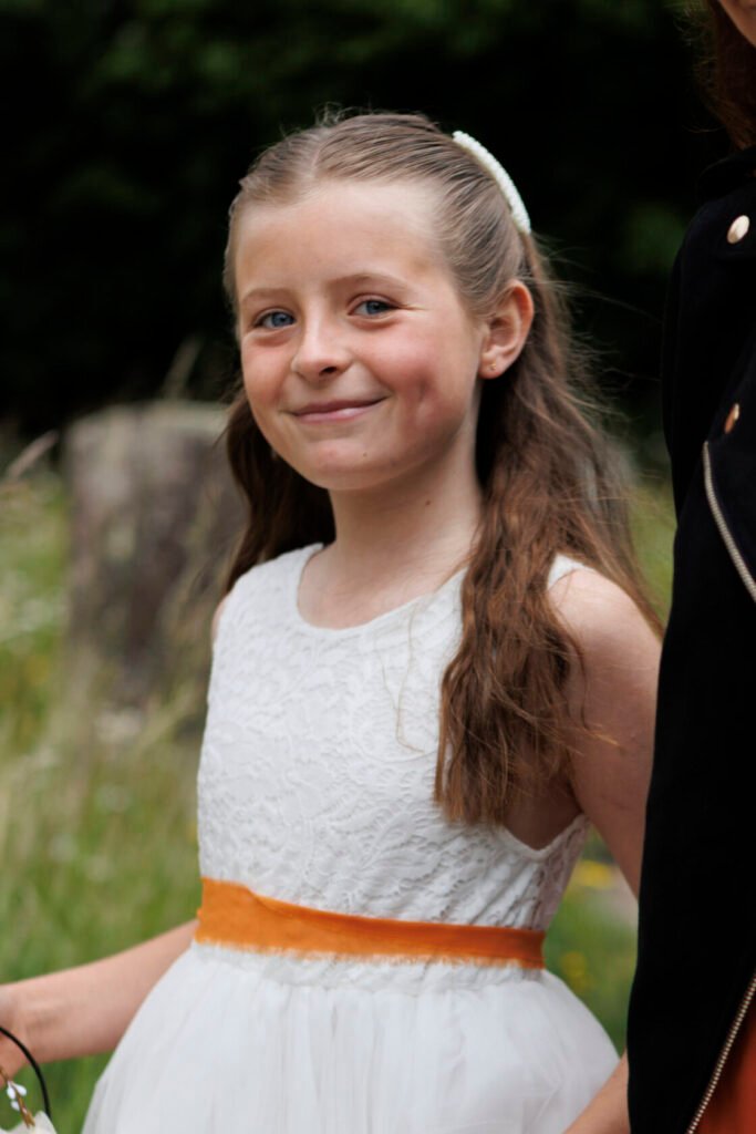 A young girl with long brown hair, wearing a white dress with lace details and an orange sash, smiles warmly while holding a small basket in a grassy outdoor setting.