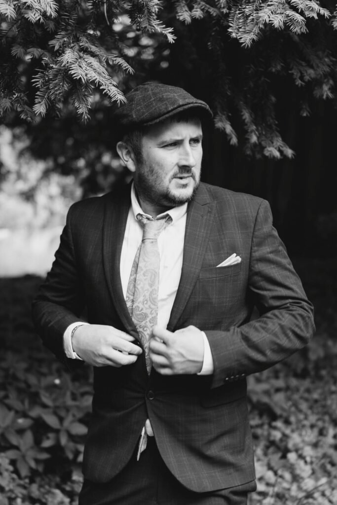 A man wearing a dark suit and patterned tie, adjusting his jacket while standing outdoors, surrounded by greenery.