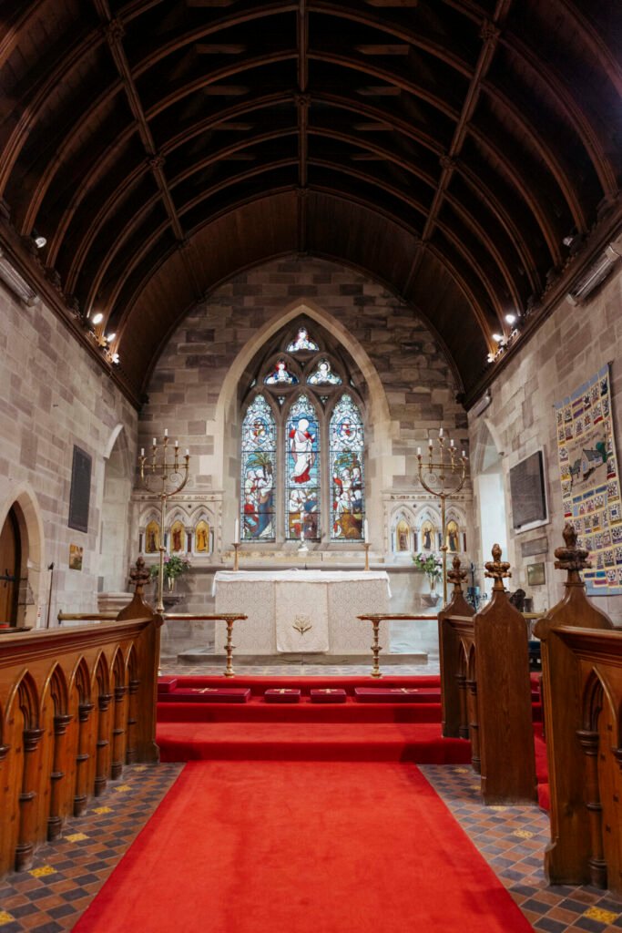 A view of the interior of a church featuring a red carpet leading to an altar with a white cloth, surrounded by intricate stained glass windows and wooden pews.