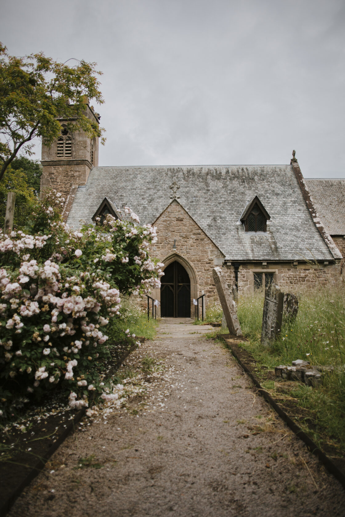 A picturesque stone church with a grey slate roof and a tall bell tower, set against a cloudy sky. The entrance is framed by lush pink roses and overgrown grass, creating a tranquil atmosphere. Shot for Lydia Turner