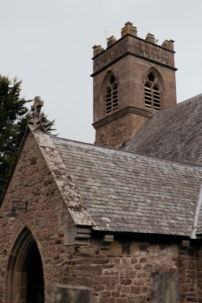 Detailed view of a historic stone church with a decorative tower and slate roof, featuring a cross on the steeple and stone carvings.