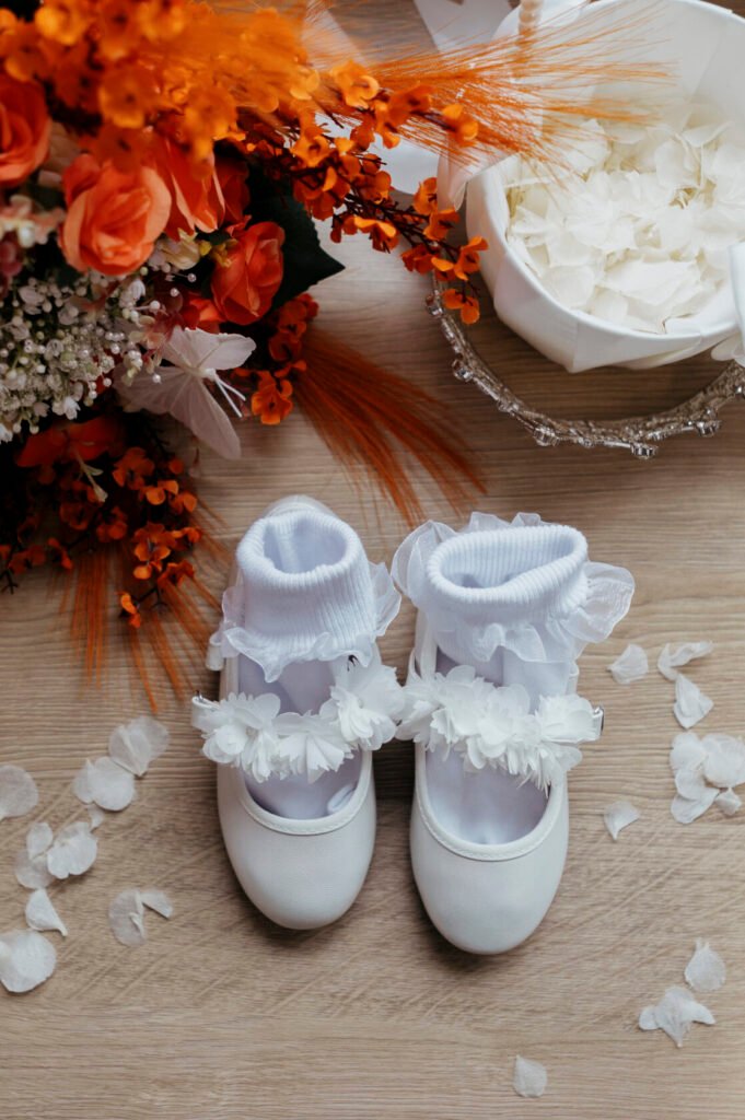 A pair of white baby shoes with fluffy white socks adorned with floral decorations, placed on a wooden surface, surrounded by flower petals and a vibrant bouquet of orange flowers.