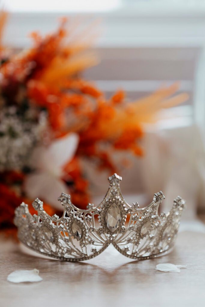 A beautifully crafted silver tiara adorned with sparkling gemstones, resting on a wooden surface with a blurred background of vibrant orange and white flowers.