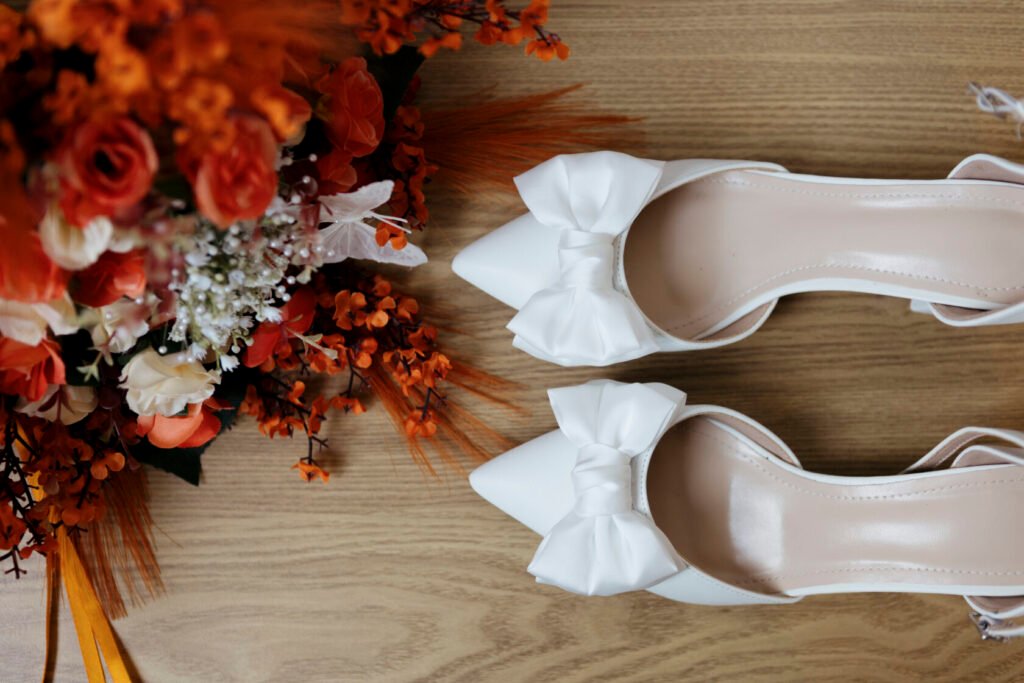 Pair of elegant white high-heeled shoes with bows, placed next to a vibrant floral bouquet featuring orange and white flowers.