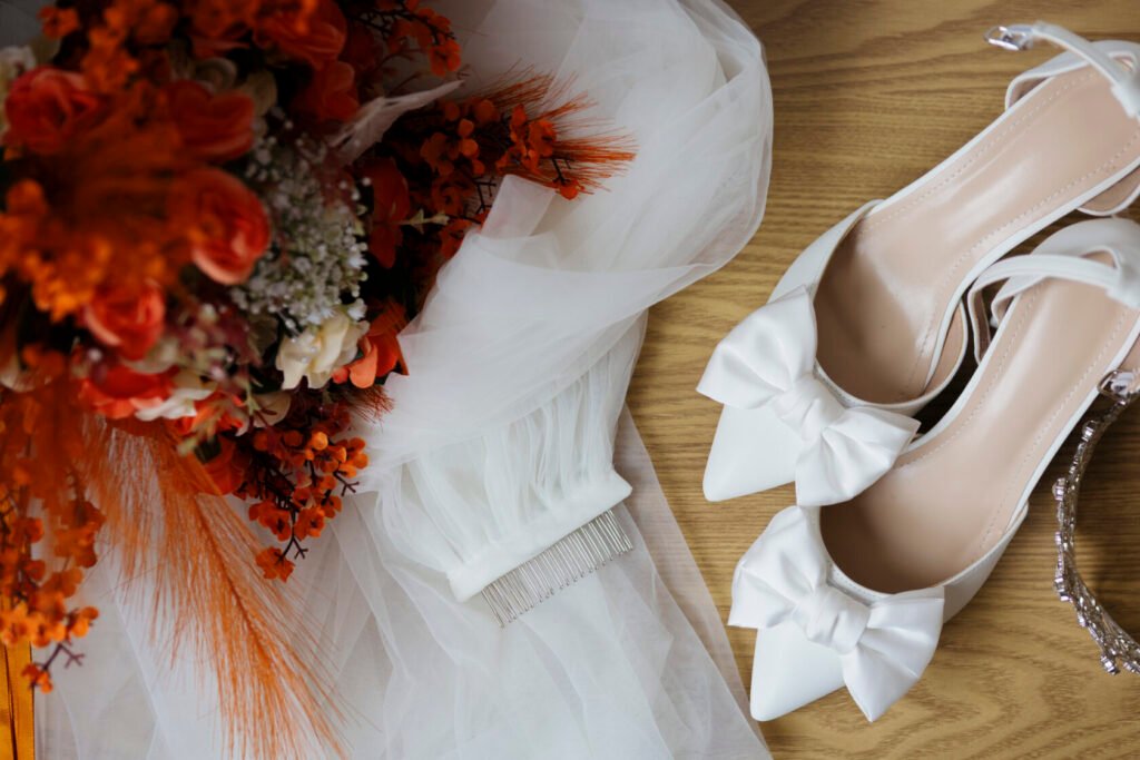A flat lay of a white bridal bouquet, white bridal shoes with bows, a delicate veil, and a hair comb on a wooden surface.