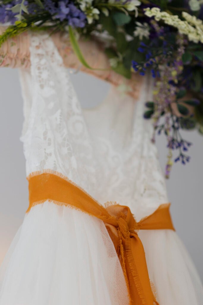 Close-up view of a lace dress featuring a frayed orange ribbon tied around the waist and floral decorations in the background.