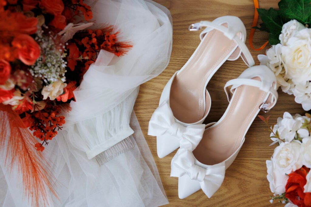 A pair of stylish white pointed-toe wedding shoes with large bows, placed beside a bridal bouquet in shades of orange and white, and a delicate white veil with a hair comb.