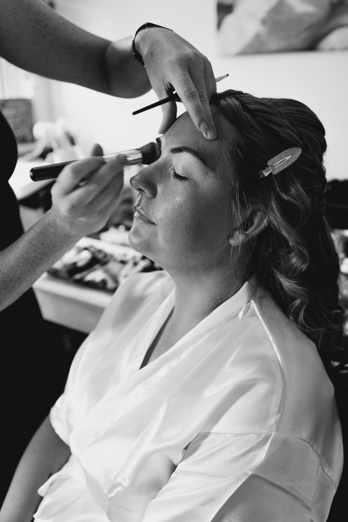 A woman sitting with her eyes closed while a makeup artist applies foundation to her face, with her hair in soft curls and wearing a white satin robe.