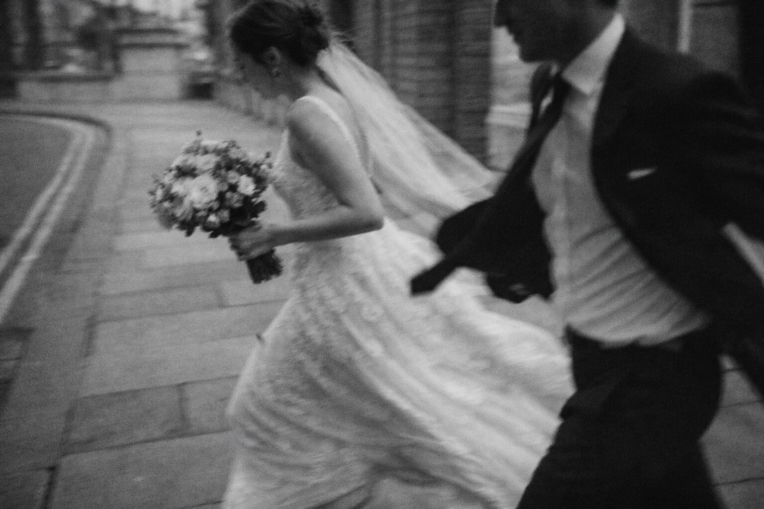 A bride in a flowing lace wedding gown runs along a cobbled street, holding a bouquet of flowers, while a man in a suit accompanies her. Shot for Lydia Turner.