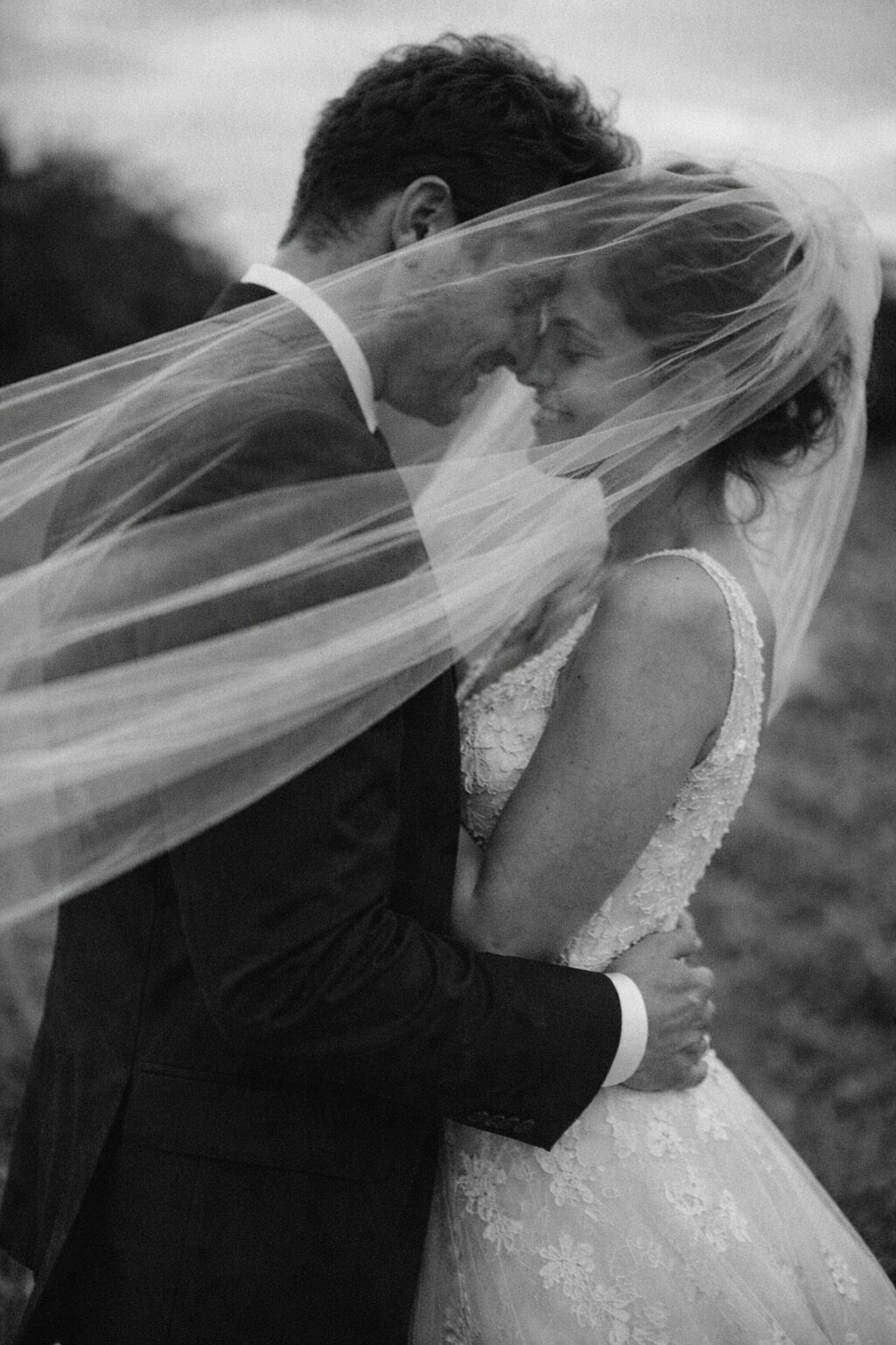 Romantic Wedding Moment Under a Veil A couple embracing closely, with a veil draped over them, sharing a tender moment. The bride wears a delicate lace gown and the groom is in a formal suit. Shot for Lydia Turner.