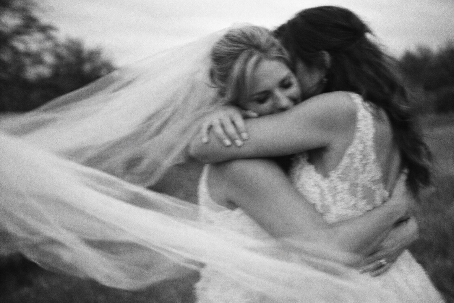 Two brides embracing warmly with flowing veils in a blurred black and white photograph. Shot for Lydia Turner.