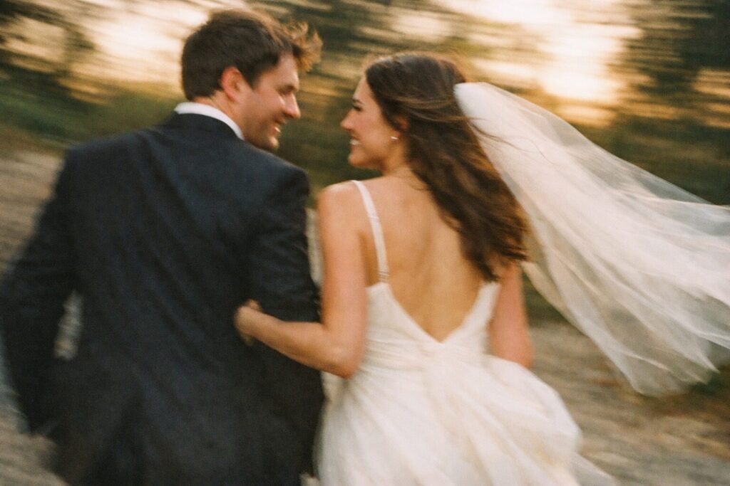 A couple joyfully running together at sunset, with the bride's veil flowing in the wind. The bride is wearing a white gown while the groom is in a black suit. Shot for Lydia Turner