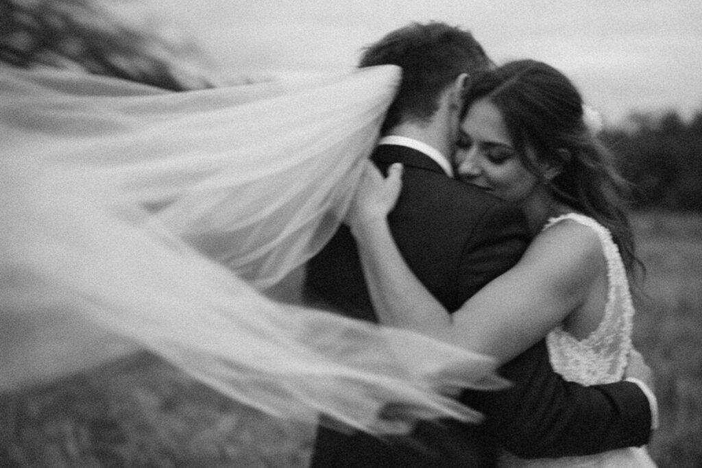 A couple tightly embracing during their wedding day, the bride's flowing veil billowing behind them. Shot for Lydia Turner
