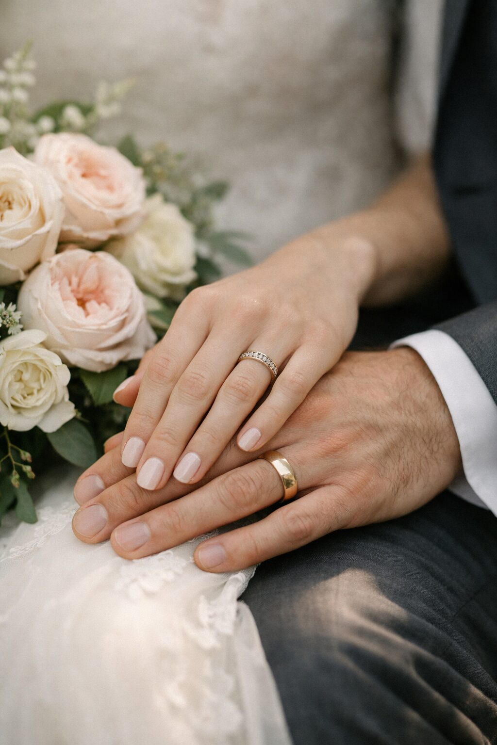 A close-up of a newlywed couple's hands with wedding rings resting on a bouquet of pale pink and white roses. Shot for Lydia Turner.