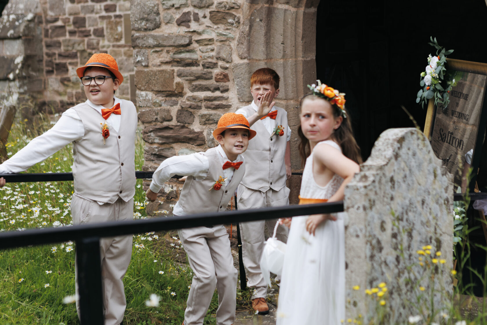 Four children dressed in wedding attire, including vests and bow ties, standing outside a building with stone walls, surrounded by flowers and greenery. One boy wears an orange hat, and a girl in a white dress has a floral crown.