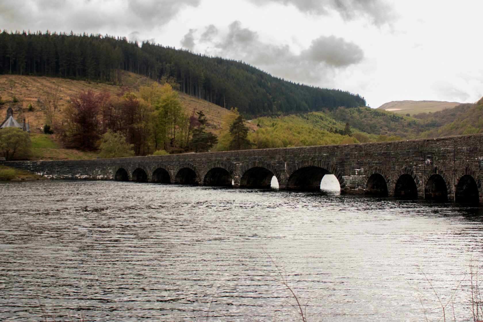 A stone bridge with multiple arches spans across a calm body of water, surrounded by lush green hills and a cloudy sky.