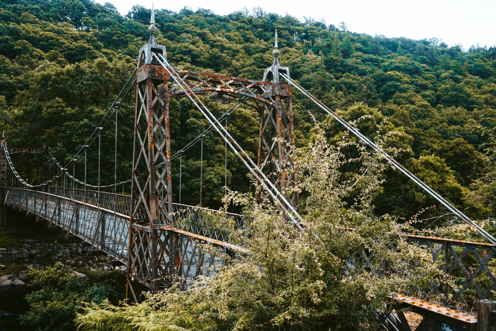 A weathered suspension bridge with rusted metal framework crossing over a river, set against a backdrop of dense green forest.