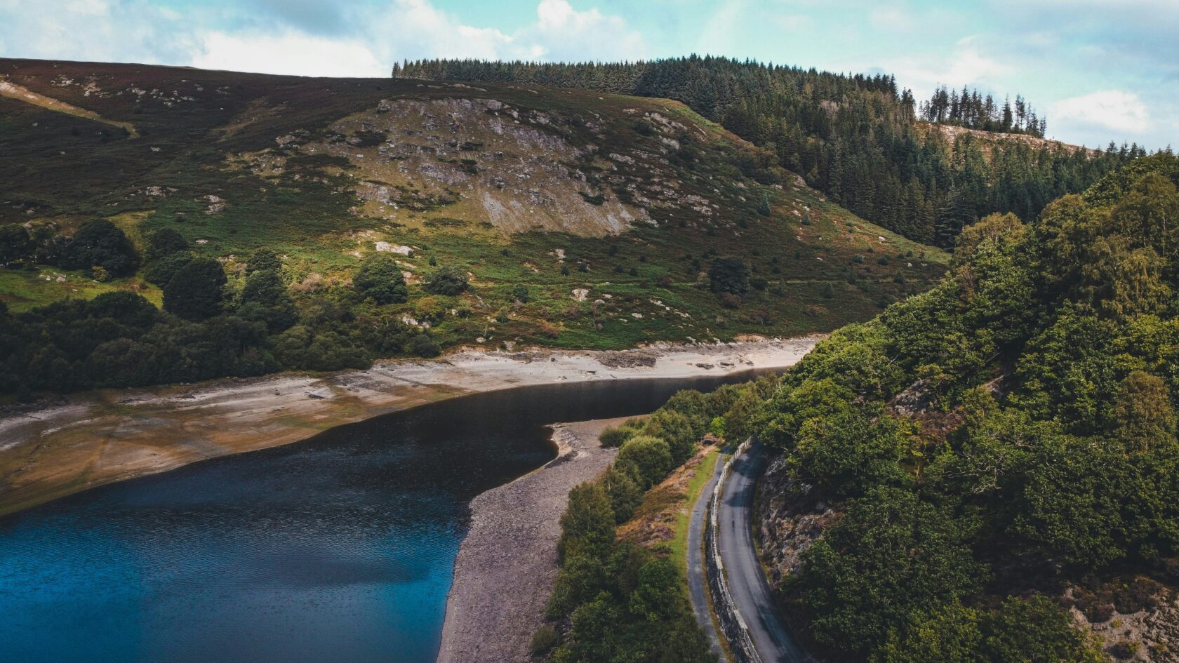 Aerial view of a tranquil reservoir surrounded by lush greenery and rolling hills, with a winding road alongside the water.