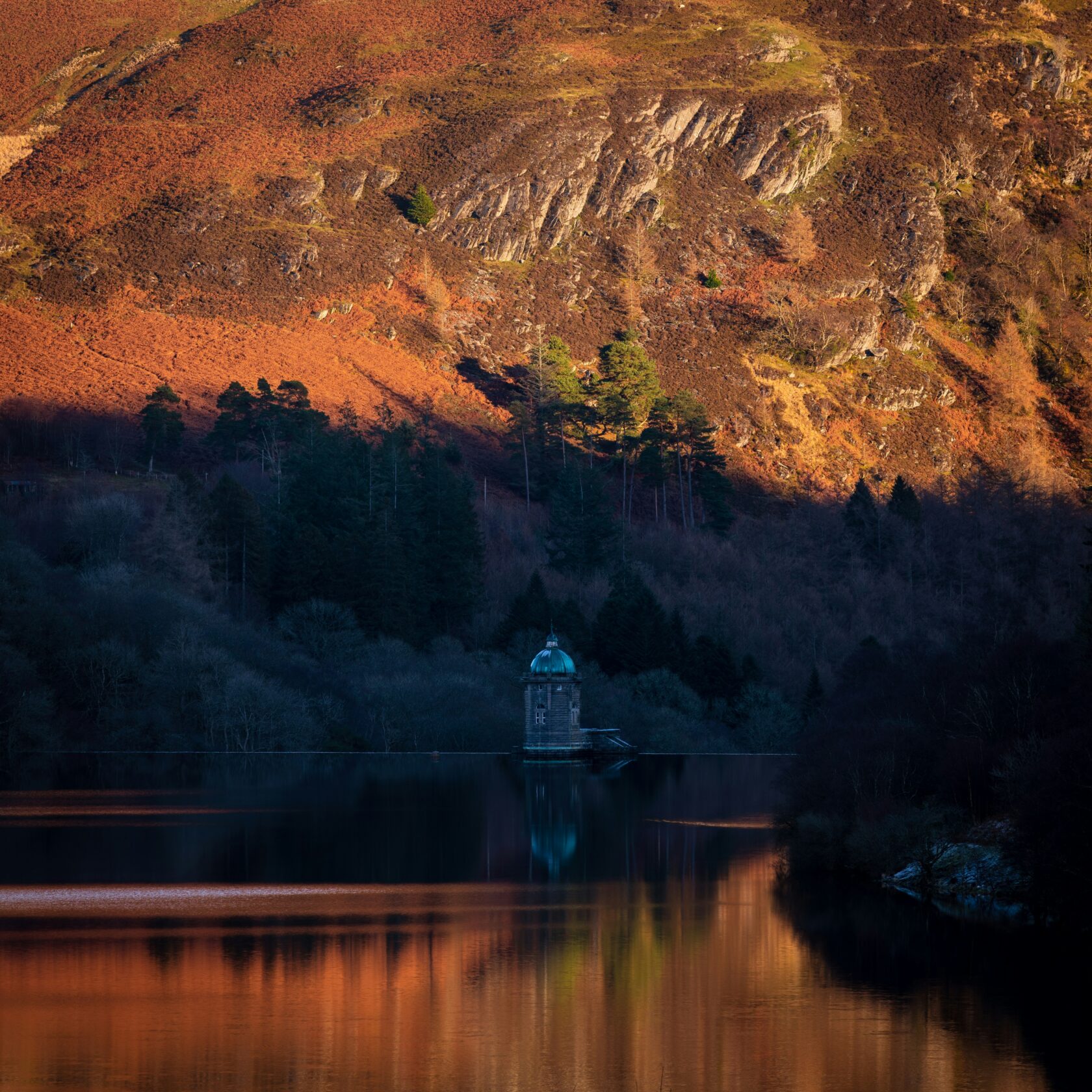 A tranquil lake reflecting the colours of the autumnal hills, with a stone structure featuring a dome in the foreground.