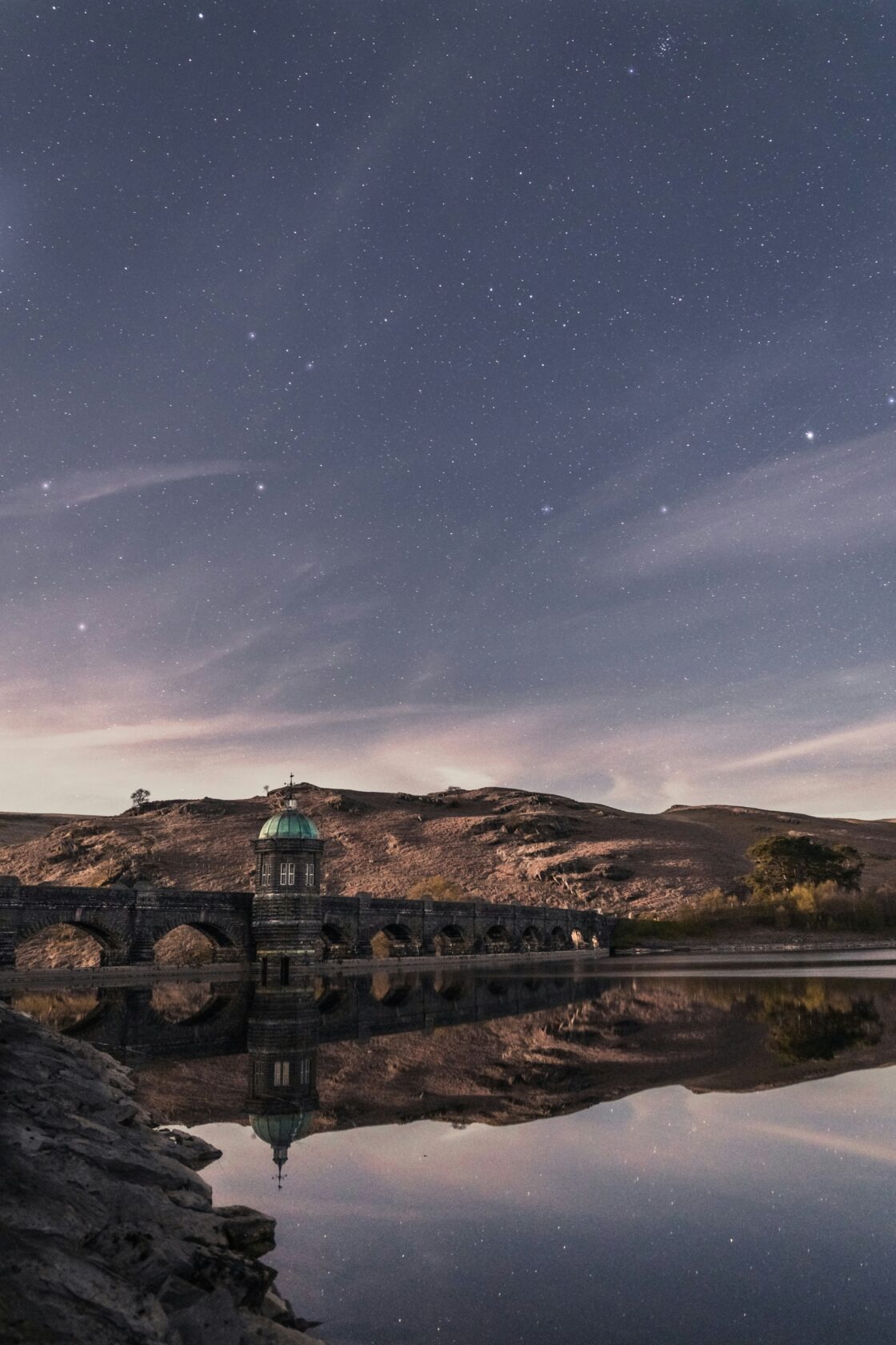 A tranquil lake reflecting a night sky filled with stars, featuring a stone structure with a green dome on the shore, surrounded by hills.