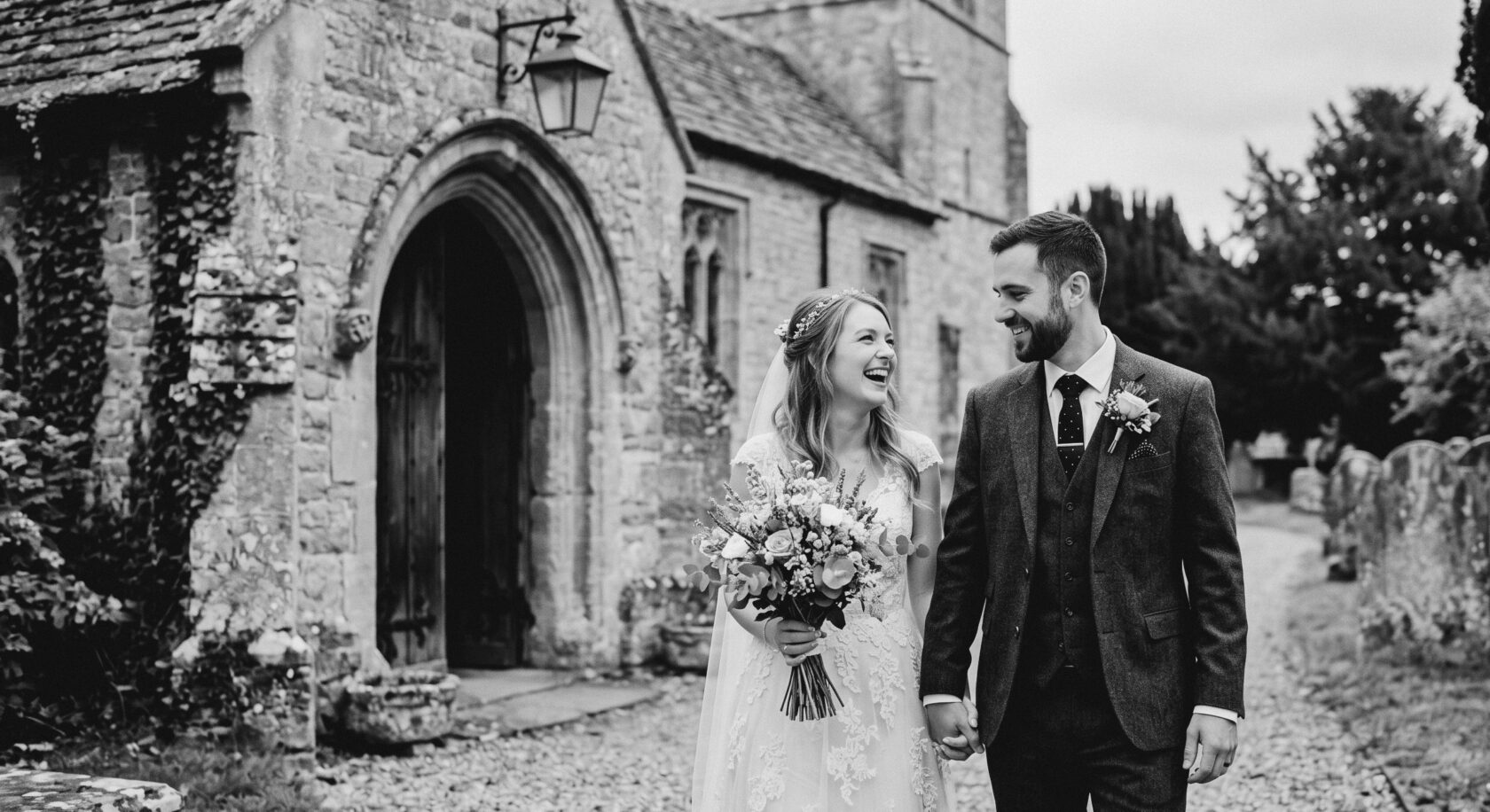 Elegant Wedding Couple Outside Historic Church A joyful bride and groom share a laugh while walking hand in hand outside a historic church. The bride wears a beautiful lace gown and holds a bouquet of flowers, while the groom is dressed in a smart suit with a boutonnière. Shot for Lydia Turner