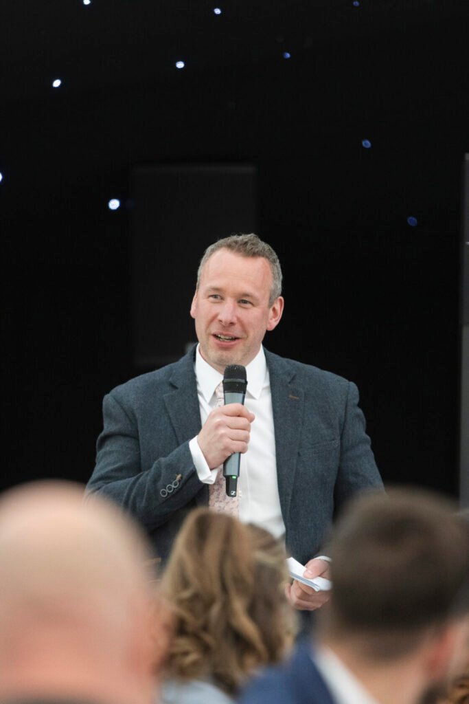 A man in a smart blazer and white shirt addresses an audience while holding a microphone, smiling and engaging. Shot for Lydia Turner.